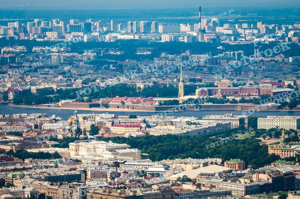 На фотографии изображен панорамный вид Санкт-Петербурга с высоты. В центре кадра располагается комплекс зданий, окружённый реками и многочисленными просторами городской застройки, в том числе и современными жилыми районами на заднем плане. Такой вид открывается с борта вертолёта или с дрона. Фотография может быть использована для туристических брошюр, исторических исследований, образовательных материалов, в новостных статьях о городе или как печатный материал в качестве украшения офисных и жилых пространств.